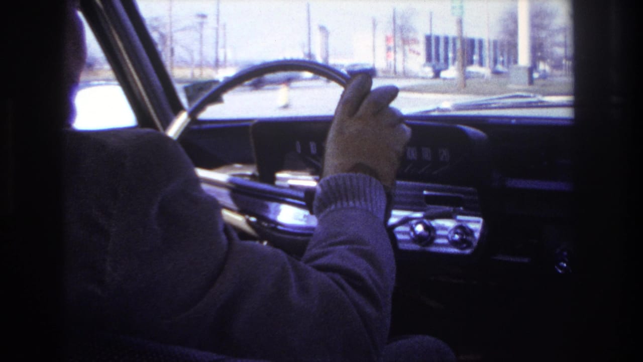 Driver's gloved hand grips the steering wheel with the dashboard and street ahead visible.