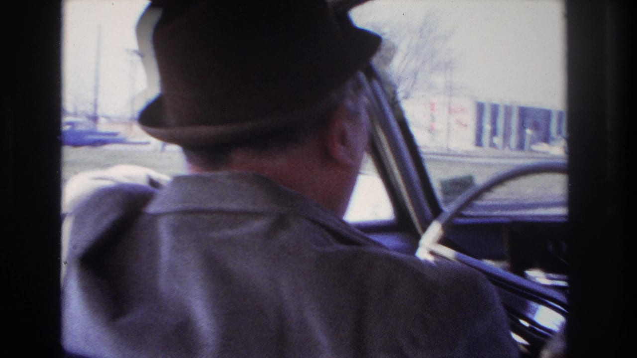 Rear-side view of the fedora-wearing driver inside the car, with the windshield and roadside buildings visible during the 1966 Hoboken drive.
