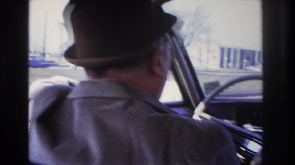Rear-side view of the fedora-wearing driver inside the car, with the windshield and roadside buildings visible during the 1966 Hoboken drive.