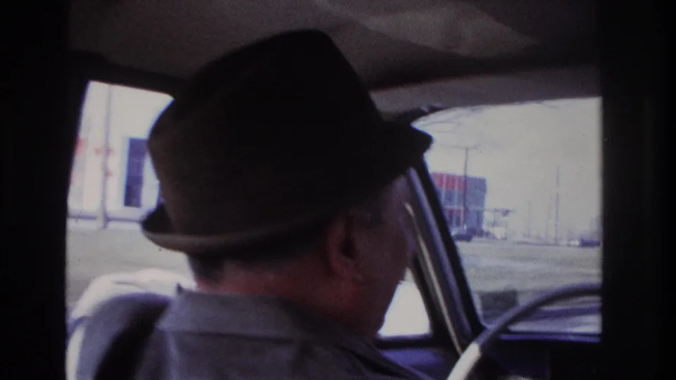 A closer rear-side view shows the fedora-wearing driver inside the car, with a bright street scene beyond the glass.