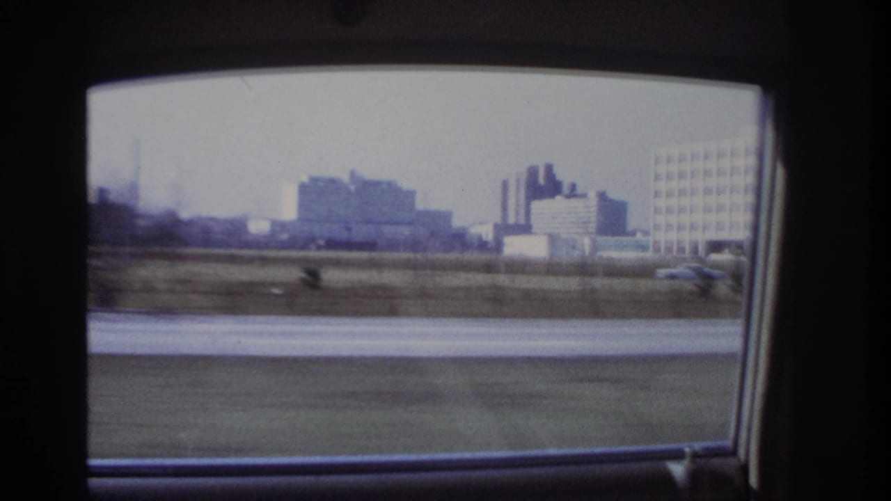 The camera turns toward the car window, showing a passing Hoboken urban landscape with low buildings, open ground, and road edge.