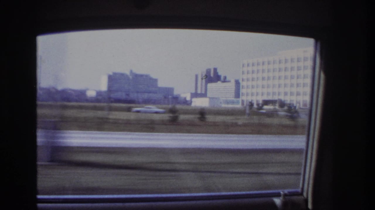 1966 Hoboken, New Jersey driving view through a car window, showing roadside grass and large grid-window buildings beyond the road.