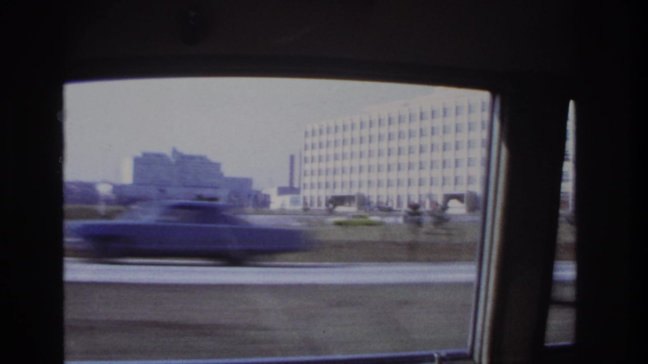 Blurred car passes outside a vehicle window with large buildings and a grassy verge behind it.