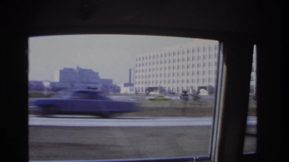 Passenger-window view in Hoboken, New Jersey, with a blurred passing car, roadside grass, and large 1960s-era buildings in the background.