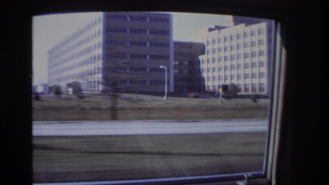 1966 Hoboken view from a moving car, showing multi-story gridded buildings, a grassy verge, and roadway blur.