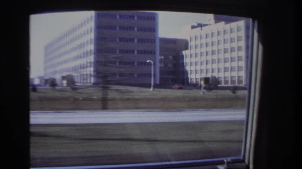 1966 Hoboken view from a moving car, showing multi-story gridded buildings, a grassy verge, and roadway blur.
