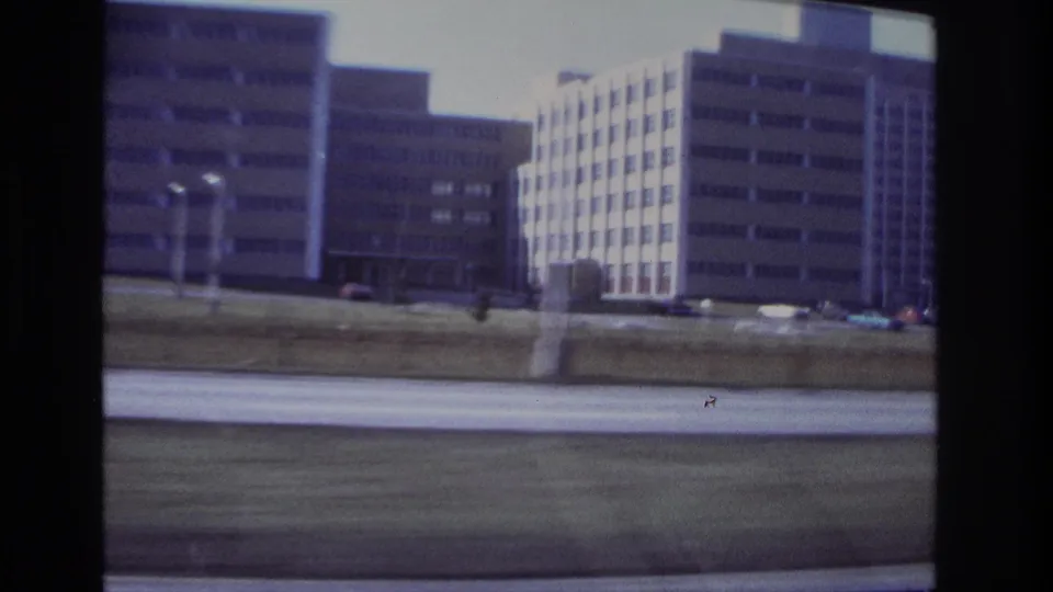 Moving-vehicle view in Hoboken, New Jersey, with gridded building facades, light poles, grass, and blurred road foreground.