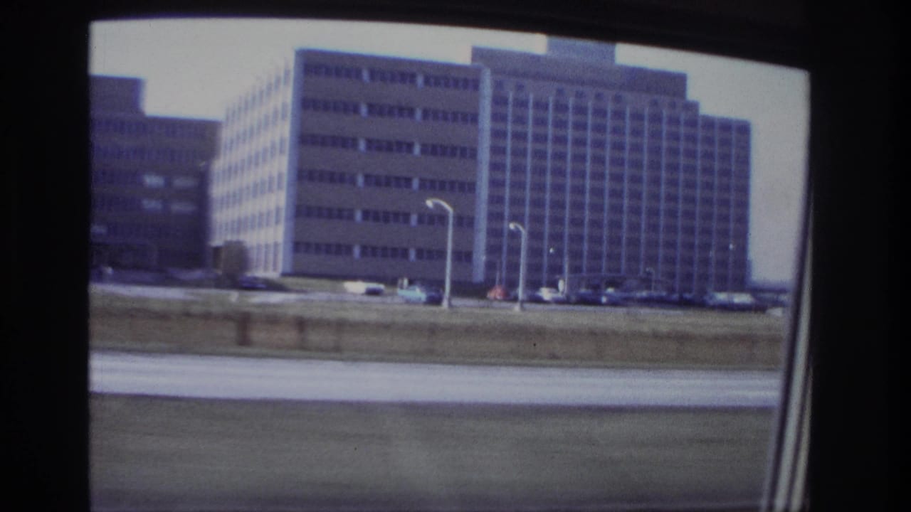 1966 Hoboken moving-car frame showing mid-century building blocks, surface parking, light poles, and a dark car-window edge.