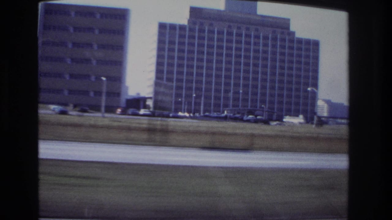 Hoboken 1966 frame with a tall grid-window building, parked cars, light poles, and roadway blur from the moving camera.