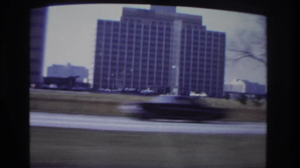 1966 Hoboken car-window view of a large gridded building with a blurred passing car in the foreground.