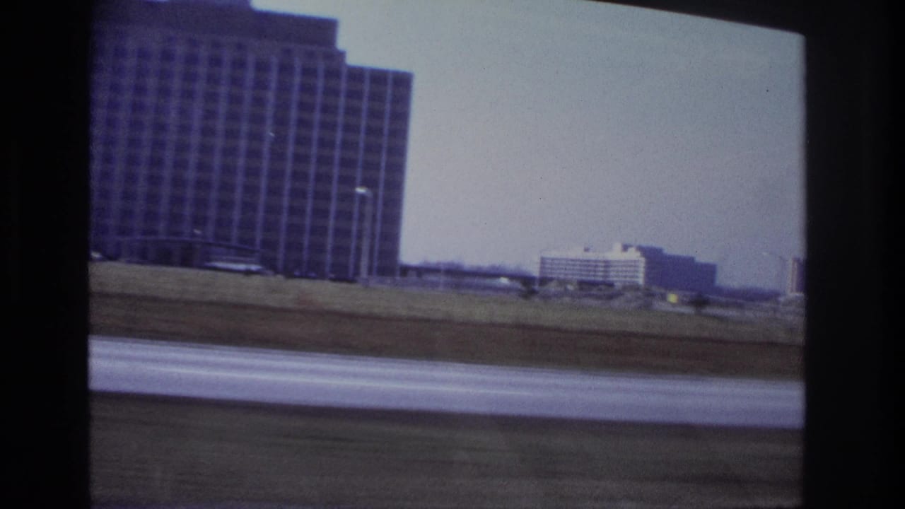 1966 Hoboken car-ride frame with a large building at left, road blur below, and low distant structures to the right.