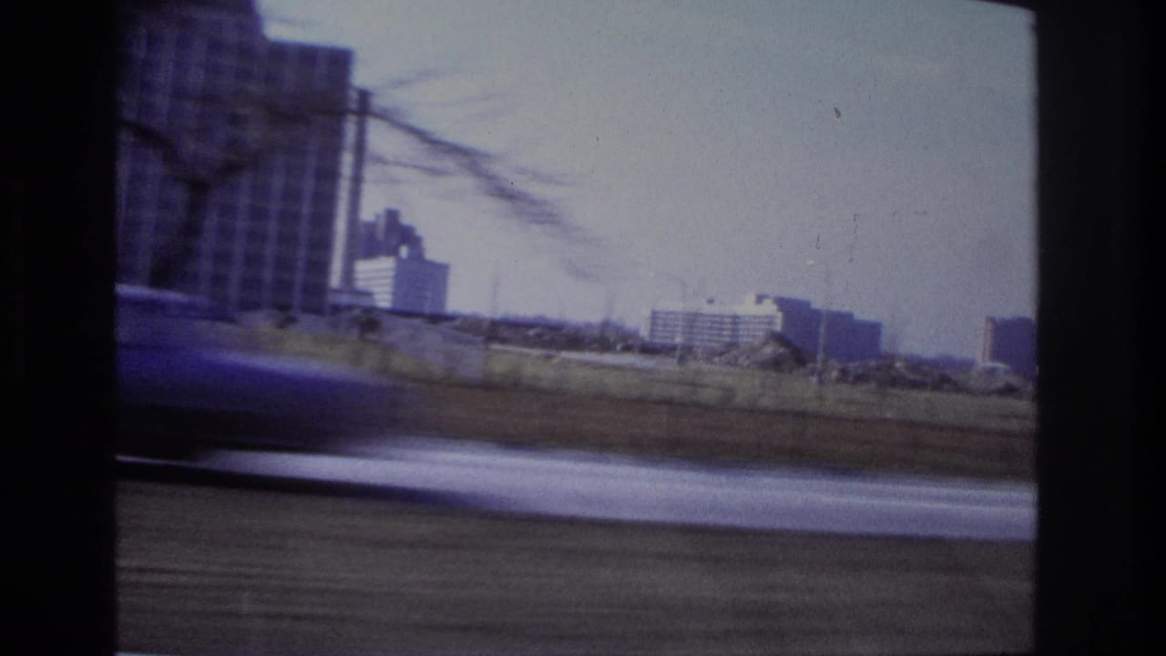 1966 Hoboken moving-car view showing distant buildings, bare branches, and a blurred vehicle or roadway foreground.