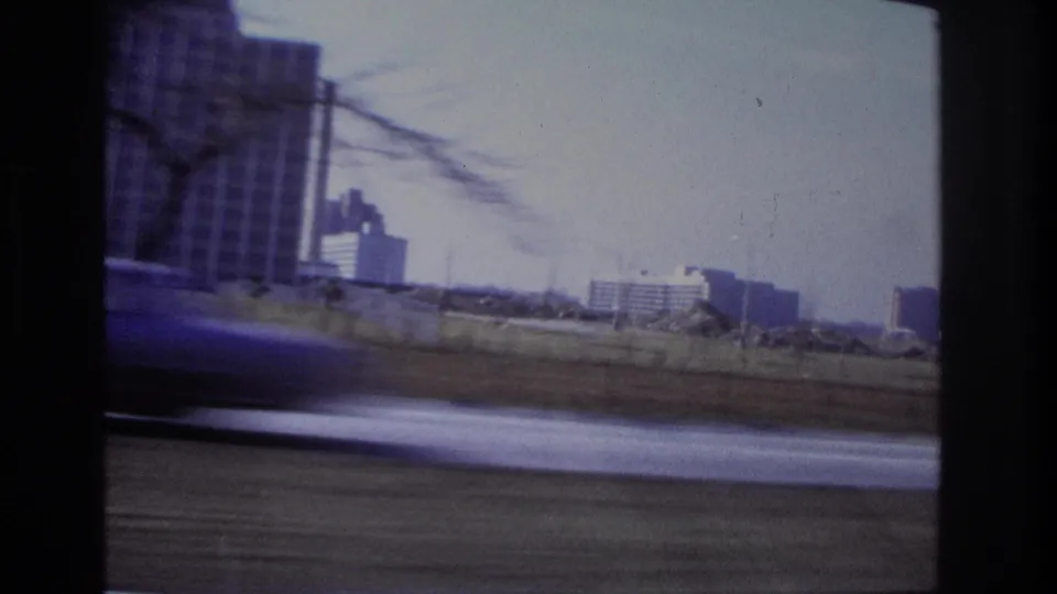 1966 Hoboken moving-car view showing distant buildings, bare branches, and a blurred vehicle or roadway foreground.