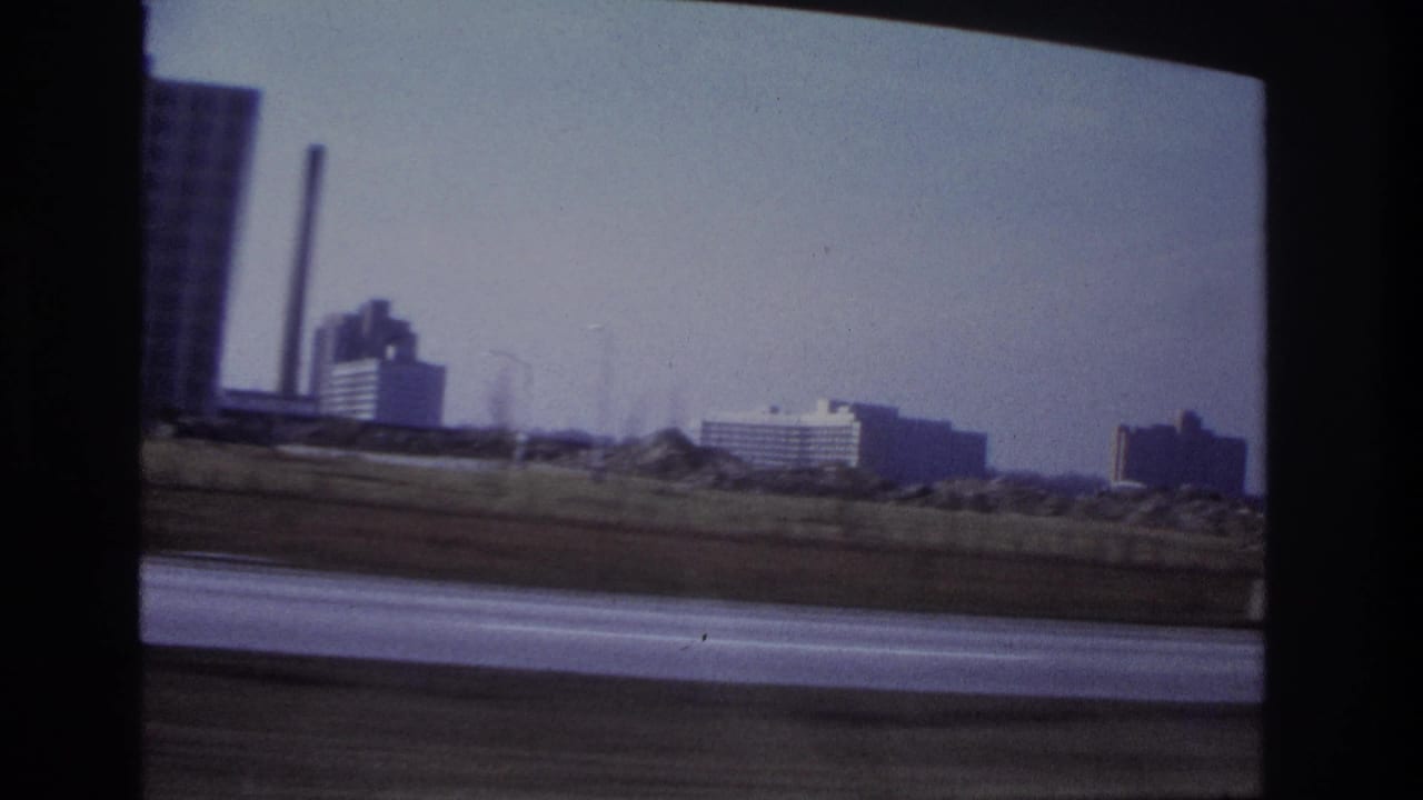 Distant buildings and industrial stacks seen past a roadside from inside a car.
