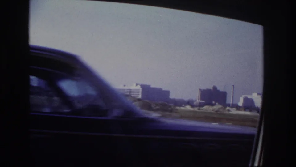A passing car blurs across a 1966 Hoboken-area road view, with distant low-rise buildings and industrial stacks beyond.