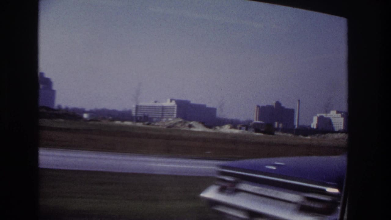 Moving-car view near Hoboken in 1966, showing distant buildings, open roadside land, and a partial car hood in the foreground.