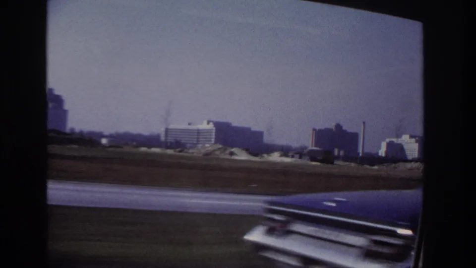 Moving-car view near Hoboken in 1966, showing distant buildings, open roadside land, and a partial car hood in the foreground.