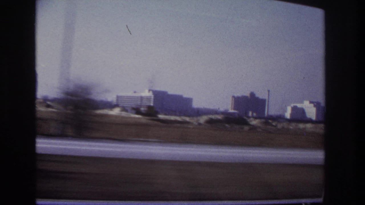 A blurred 1966 car-window view near Hoboken with roadside motion, distant buildings, industrial stacks, and a pale hazy sky.