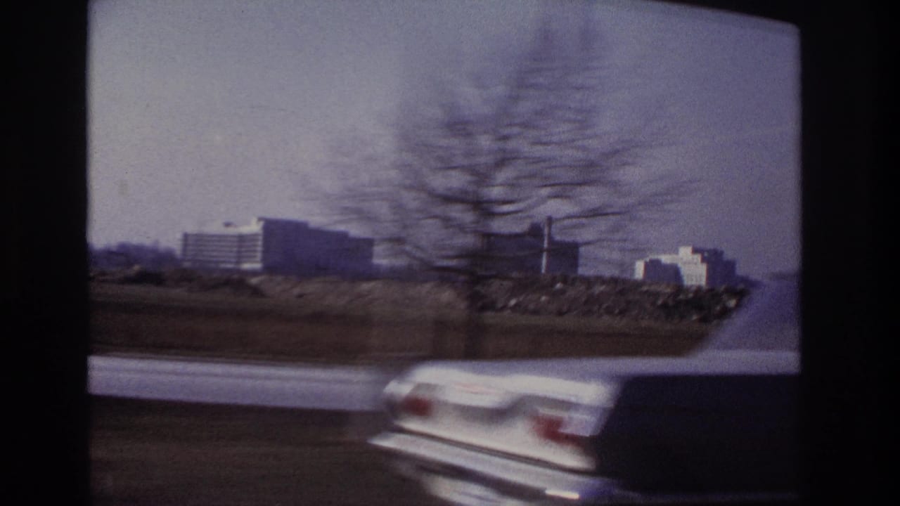 A blurred white car passes in front of a 1966 Hoboken highway view with distant buildings and roadside land.