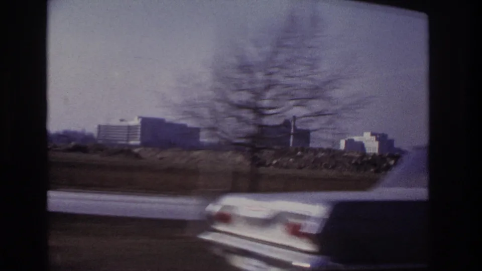 A blurred white car passes in front of a 1966 Hoboken highway view with distant buildings and roadside land.