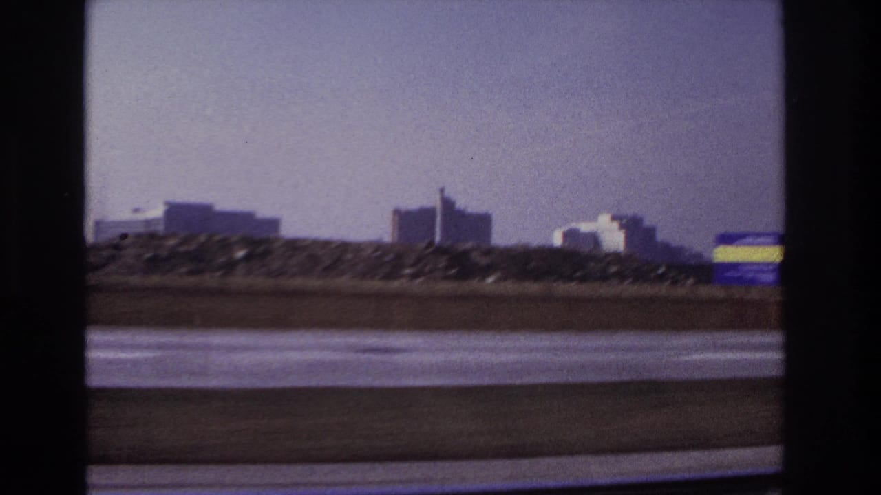 1966 Hoboken-area 8mm frame from a moving vehicle, showing a highway barrier, distant buildings, open sky, and motion blur.