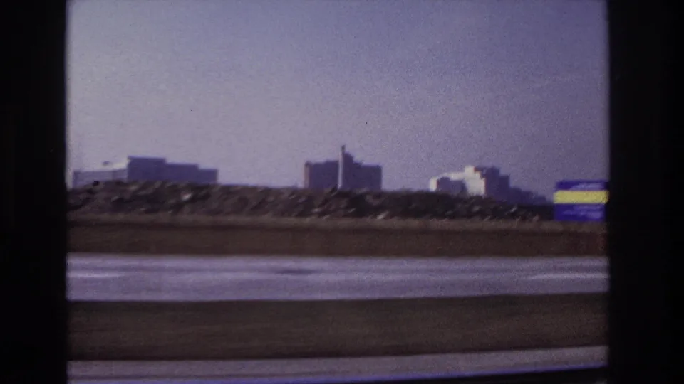 1966 Hoboken-area 8mm frame from a moving vehicle, showing a highway barrier, distant buildings, open sky, and motion blur.