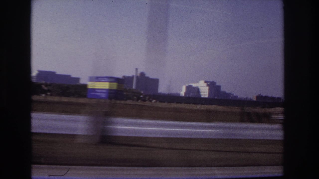 1966 Hoboken-area road footage showing a blurred vehicle crossing the foreground with buildings and a smokestack in the distance.