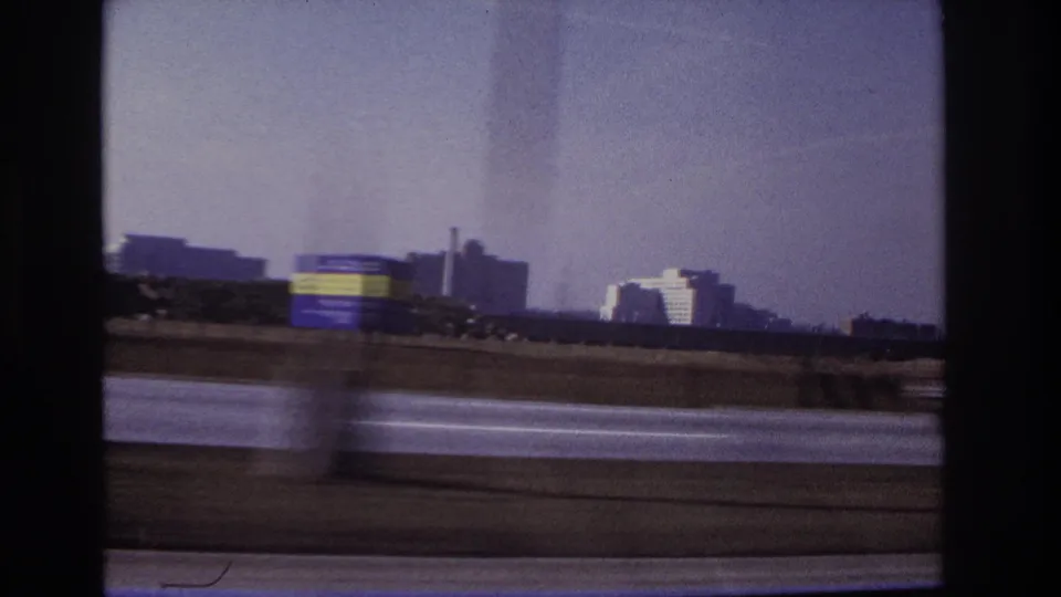 1966 Hoboken-area road footage showing a blurred vehicle crossing the foreground with buildings and a smokestack in the distance.