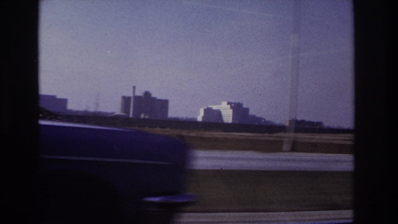 1966 Hoboken-area 8mm frame with a dark blurred car in the foreground and industrial-looking buildings beyond the road.