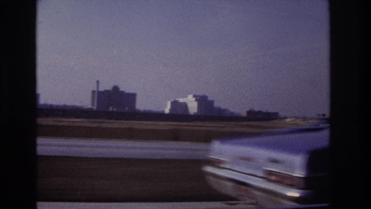 1966 Hoboken-area highway frame with a light-colored car blurred in the foreground and distant buildings under a clear sky.