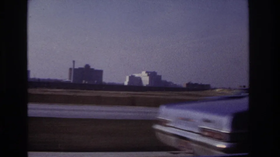 1966 Hoboken-area highway frame with a light-colored car blurred in the foreground and distant buildings under a clear sky.