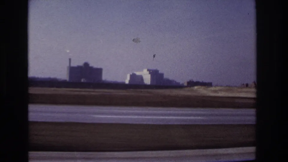 1966 Hoboken-area home-movie view through a car window, with roadway blur and distant industrial buildings on the horizon.