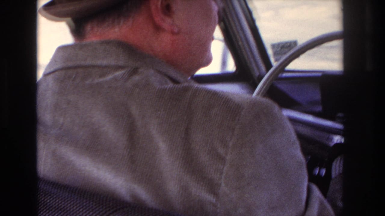 Man in a hat and corduroy jacket driving an older car, seen from behind.