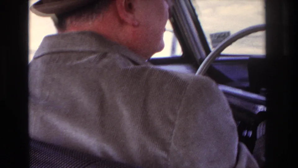 Man driving an older car near Hoboken in 1966, filmed from the rear passenger area with the steering wheel and window visible.