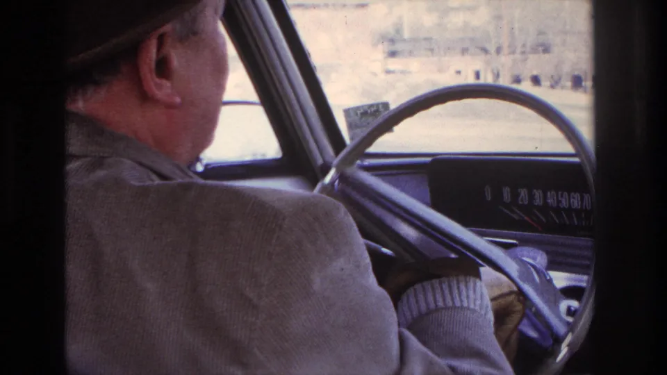 Rear-seat view of a man in a hat and jacket driving an older car, with dashboard details and bright exterior scenery ahead.