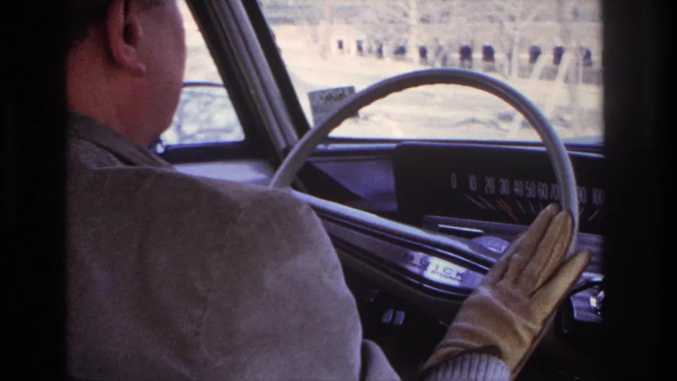 Inside a 1966 Hoboken-area driving shot, the driver’s gloved hand reaches near the older car dashboard and analog speedometer.