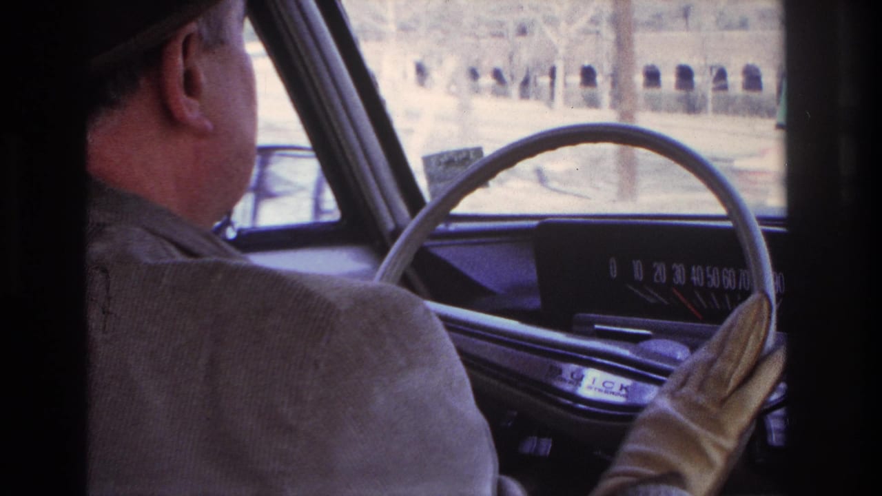 In 1966 Hoboken home-movie footage, a man in a dark fedora drives while the steering wheel, dashboard, and passing exterior are visible.