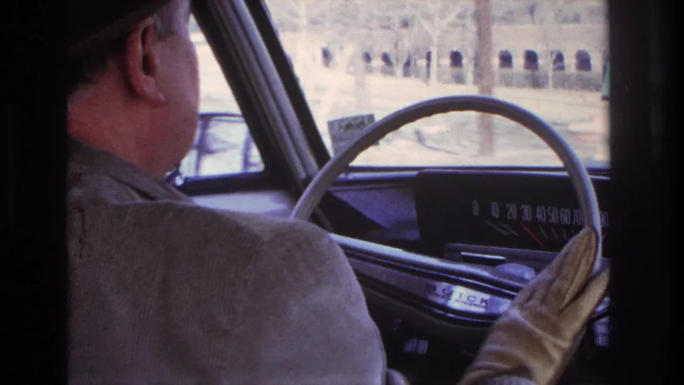 In 1966 Hoboken home-movie footage, a man in a dark fedora drives while the steering wheel, dashboard, and passing exterior are visible.