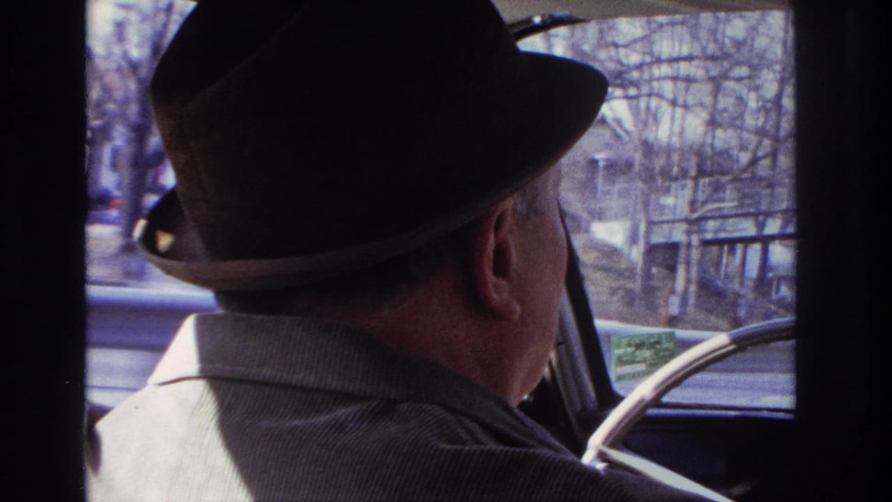 A man in a fedora drives through Hoboken in 1966, seen beside the steering wheel with street scenery through the front glass.