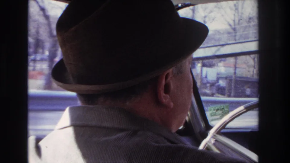 1966 Hoboken-area home-movie frame showing a man in a dark fedora and corduroy jacket driving, with bare trees outside the window.