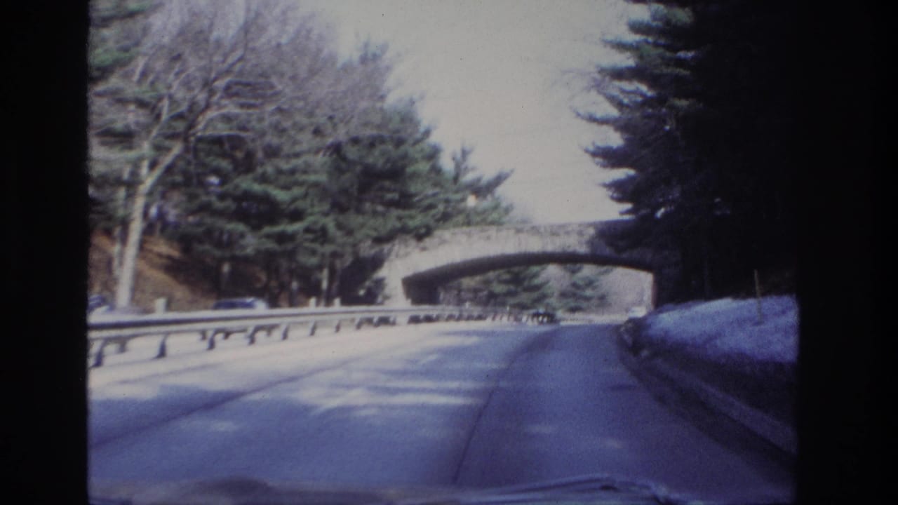 Forward-looking 8mm frame from inside a car showing a tree-lined roadway, guardrail, and stone arch bridge ahead.