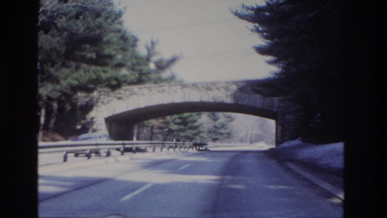 1966 8mm road-trip view near Hoboken, New Jersey, approaching a stone arch bridge on a wooded winter roadway.