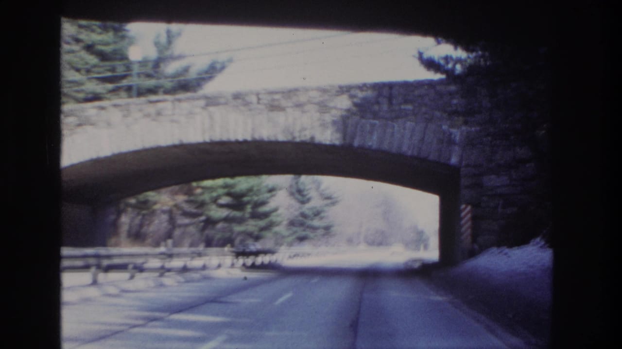 Closer windshield view of a stone arch bridge spanning a road bordered by trees and snow.