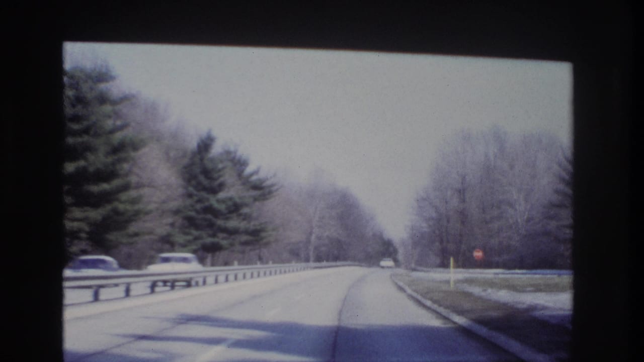 1966 8mm-style forward driving view on a wooded New Jersey roadway, with guardrails, bare trees, and cars ahead.
