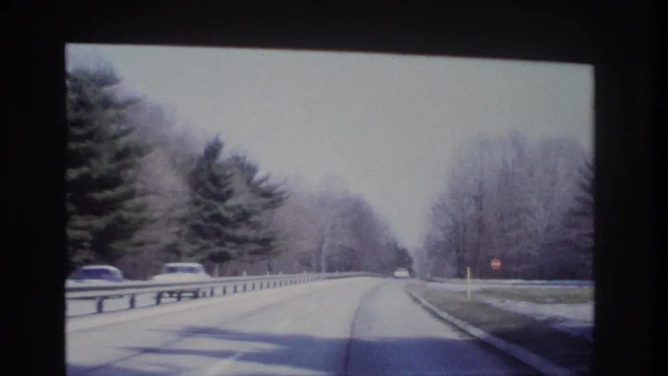 1966 8mm-style forward driving view on a wooded New Jersey roadway, with guardrails, bare trees, and cars ahead.