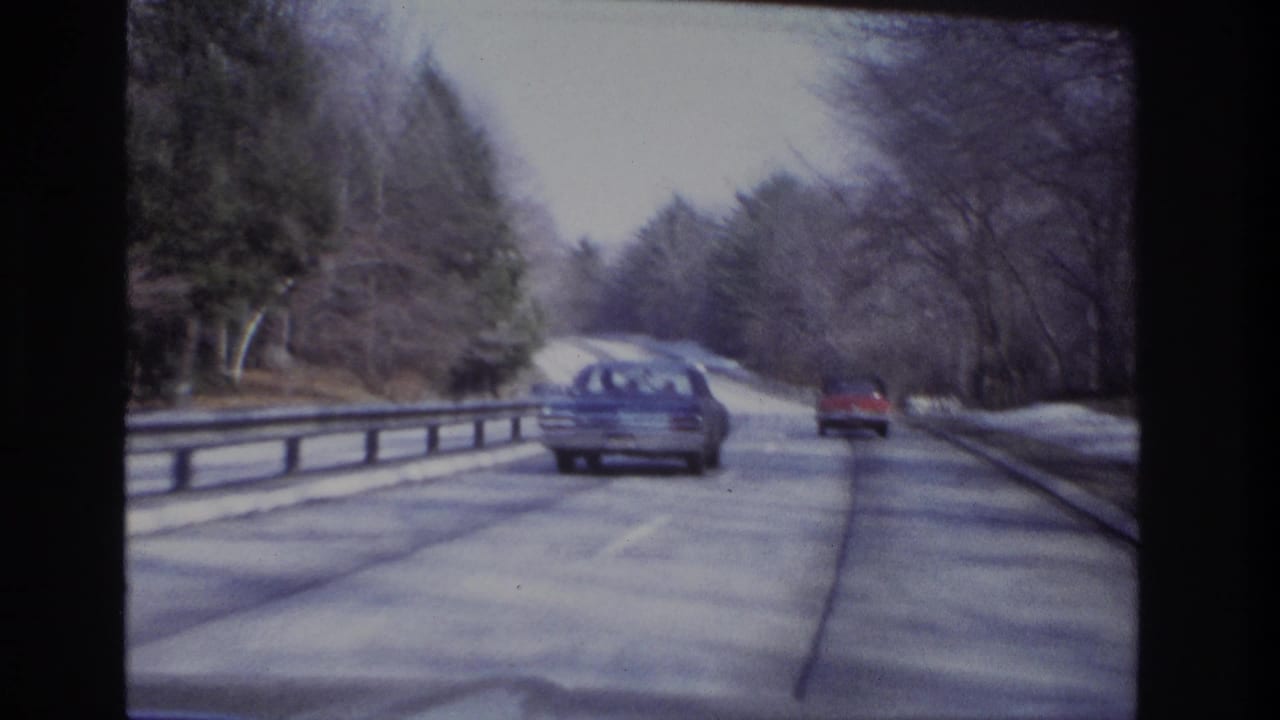 A blue sedan and red car travel ahead on a wooded New Jersey road in 1966 home-movie highway footage.