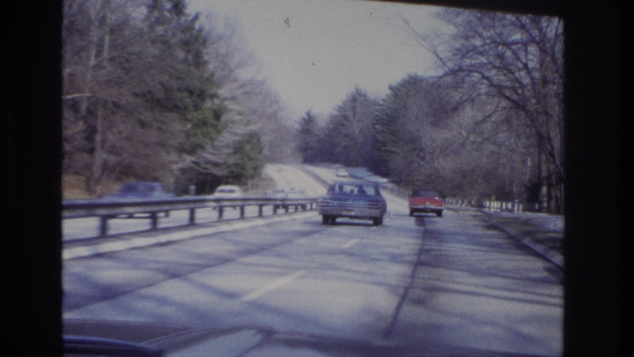 1966 home-movie frame of cars ahead on a tree-lined New Jersey roadway with a guardrail and muted winter light.