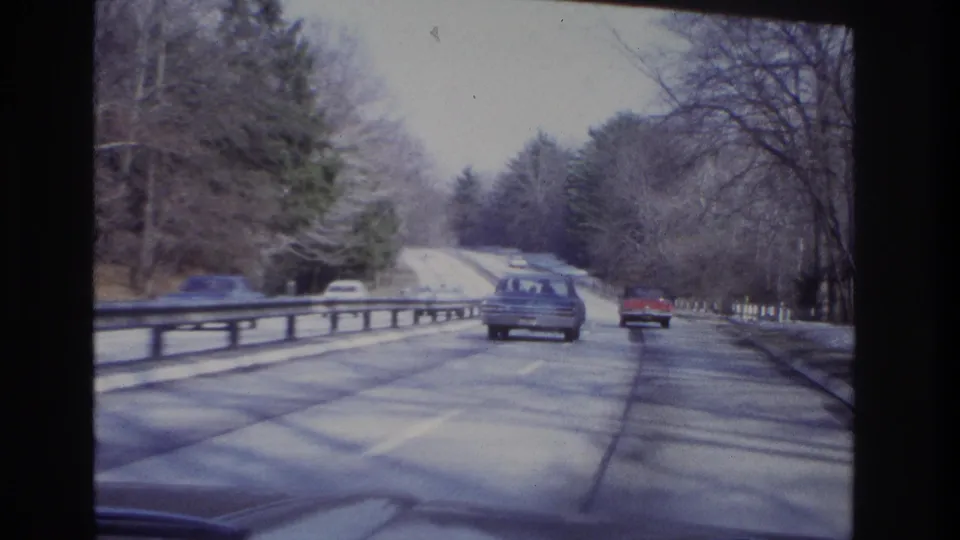 1966 home-movie frame of cars ahead on a tree-lined New Jersey roadway with a guardrail and muted winter light.
