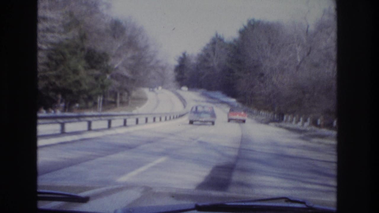 1966 8mm-style view from inside a moving vehicle on a winding road with classic cars, bare trees, and snow near the shoulder.