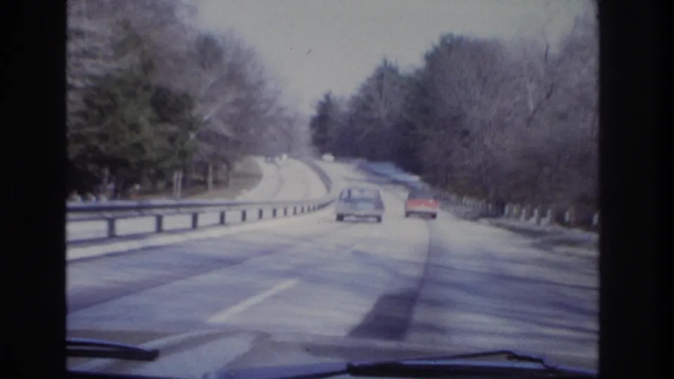 1966 8mm-style view from inside a moving vehicle on a winding road with classic cars, bare trees, and snow near the shoulder.